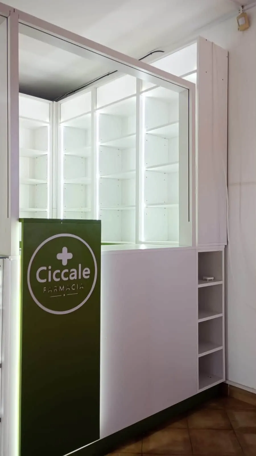 Columna con el logo circular de Farmacia Ciccale sobre frente verde, junto a estanterías vidriadas e iluminadas.