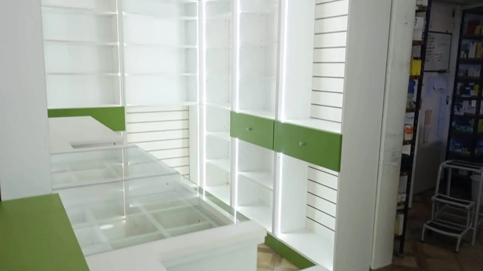 Mostrador con tapa de vidrio y cajoneras, estantería de slatwall con estantes blancos e iluminación LED, y acceso al depósito.