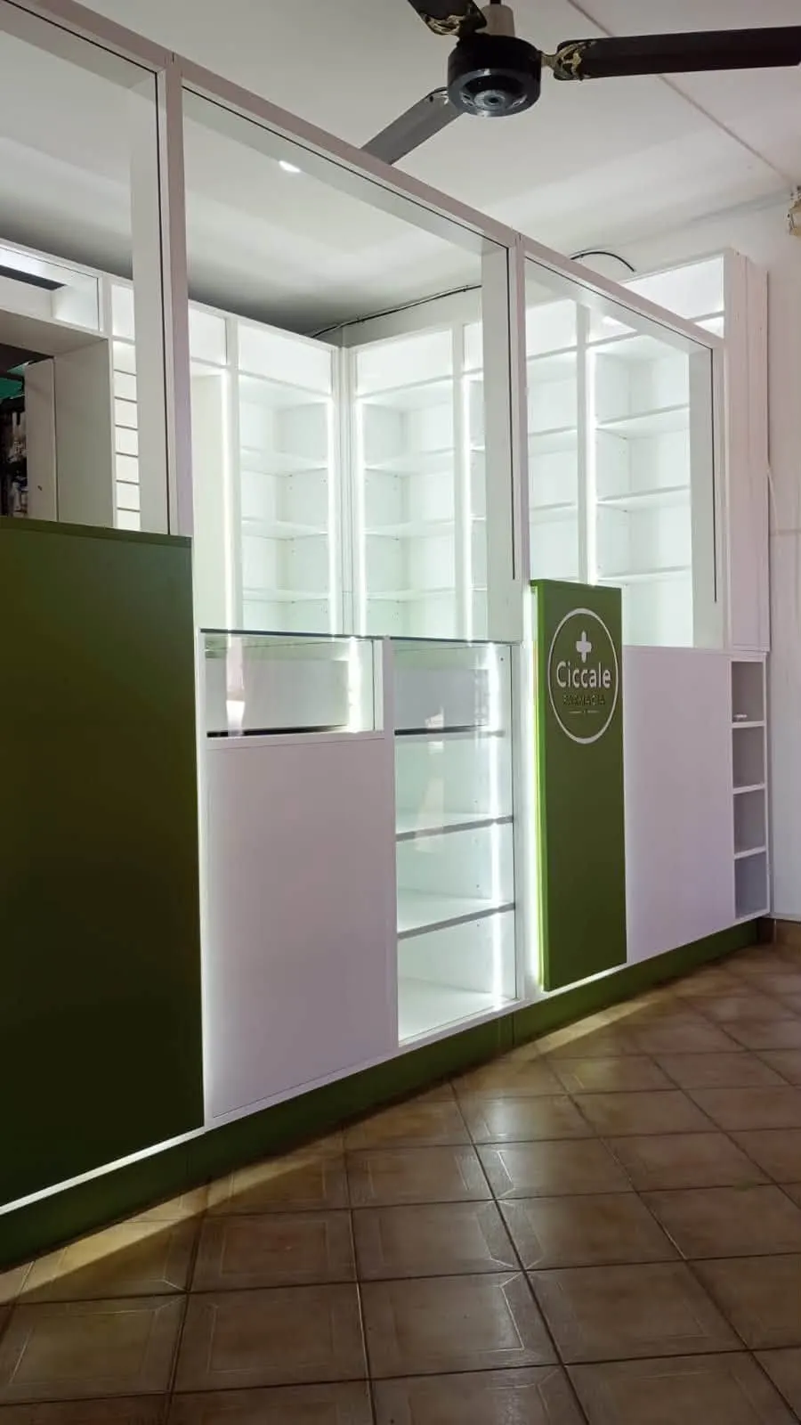 Mostrador principal de Farmacia Ciccale con frente verde, columna señalizadora con el logo y estantería alta vidriada e iluminada.