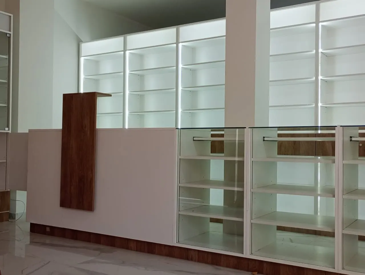 Mostrador de atención en melamina blanca con friso superior en madera y estanterías perimetrales de farmacia recién instaladas.