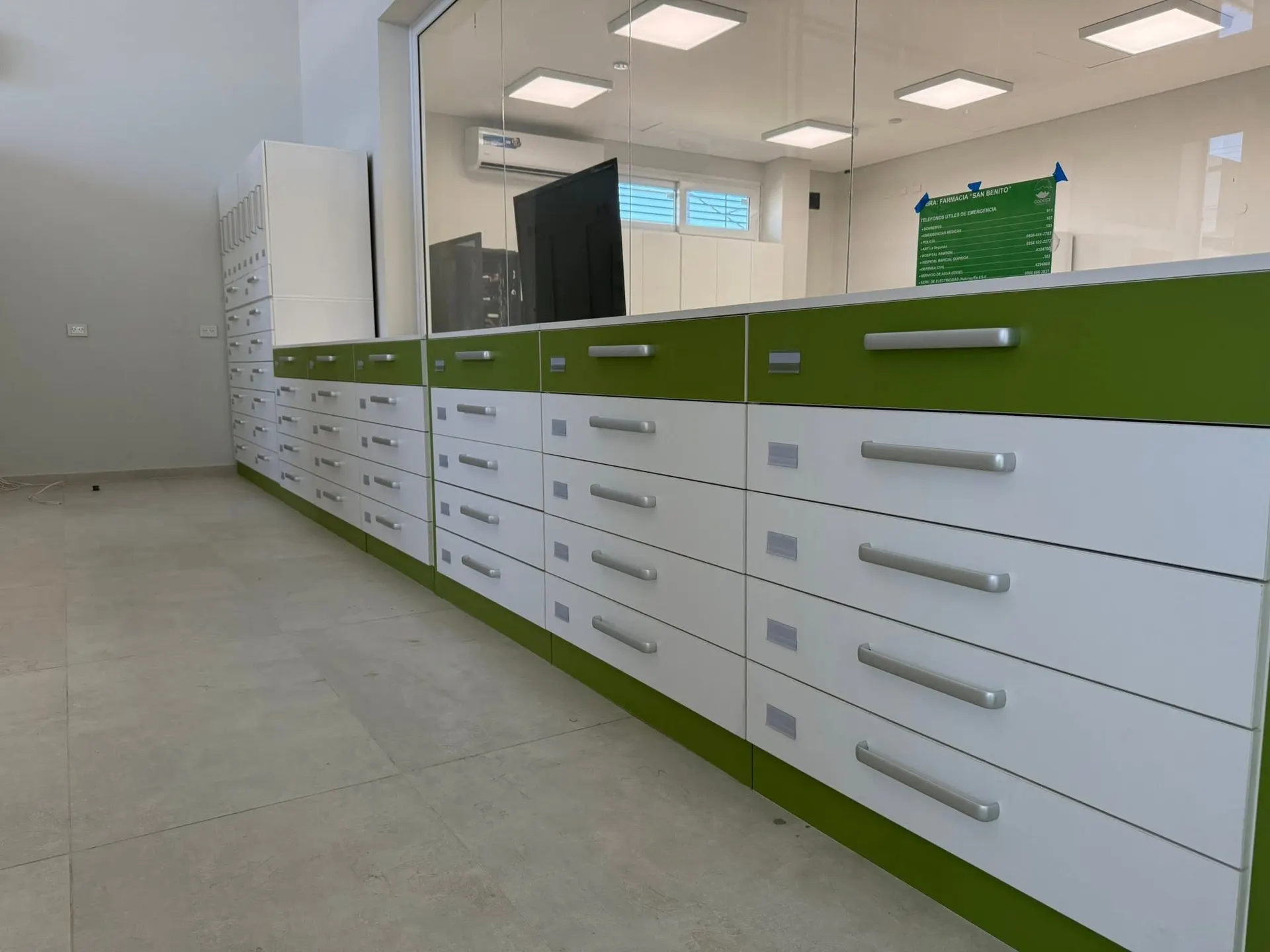 Mostrador perimetral en blanco y verde con cajoneras y estanterías altas en la obra de Farmacia San Benito.