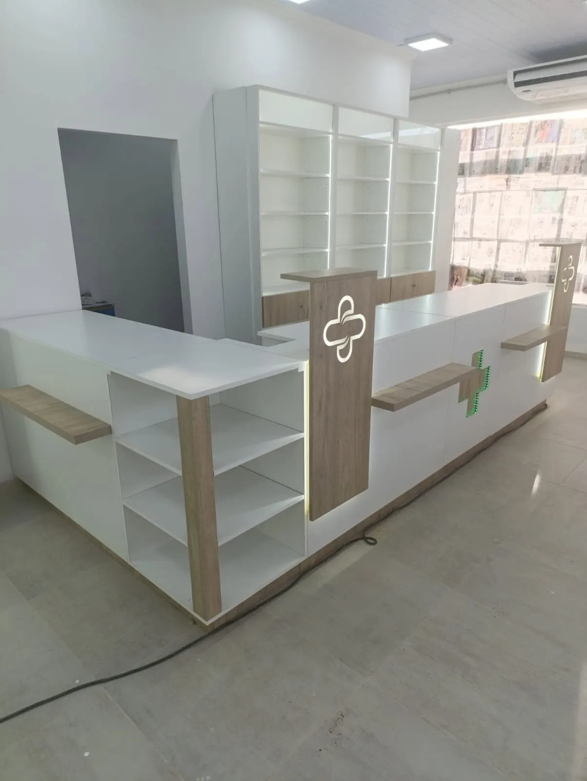Mostrador de atención con cruz farmacéutica retroiluminada en el frente y estantes laterales.