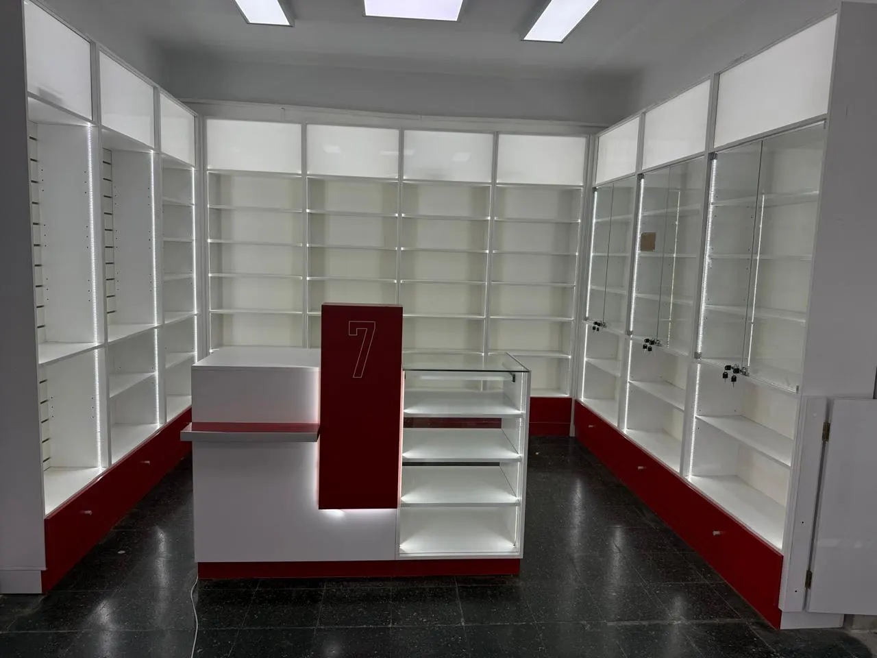 Salón de venta con puesto de atención numerado, mostradores de vidrio y estantería perimetral iluminada.