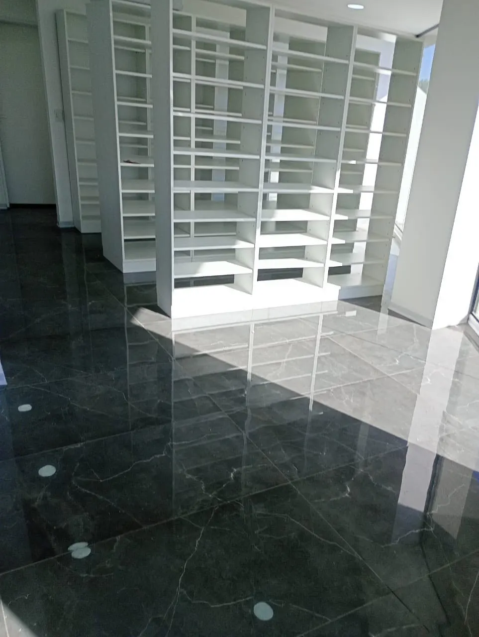 Torre de estanterías blancas reflejada en piso de mármol, junto a un pasillo de circulación.